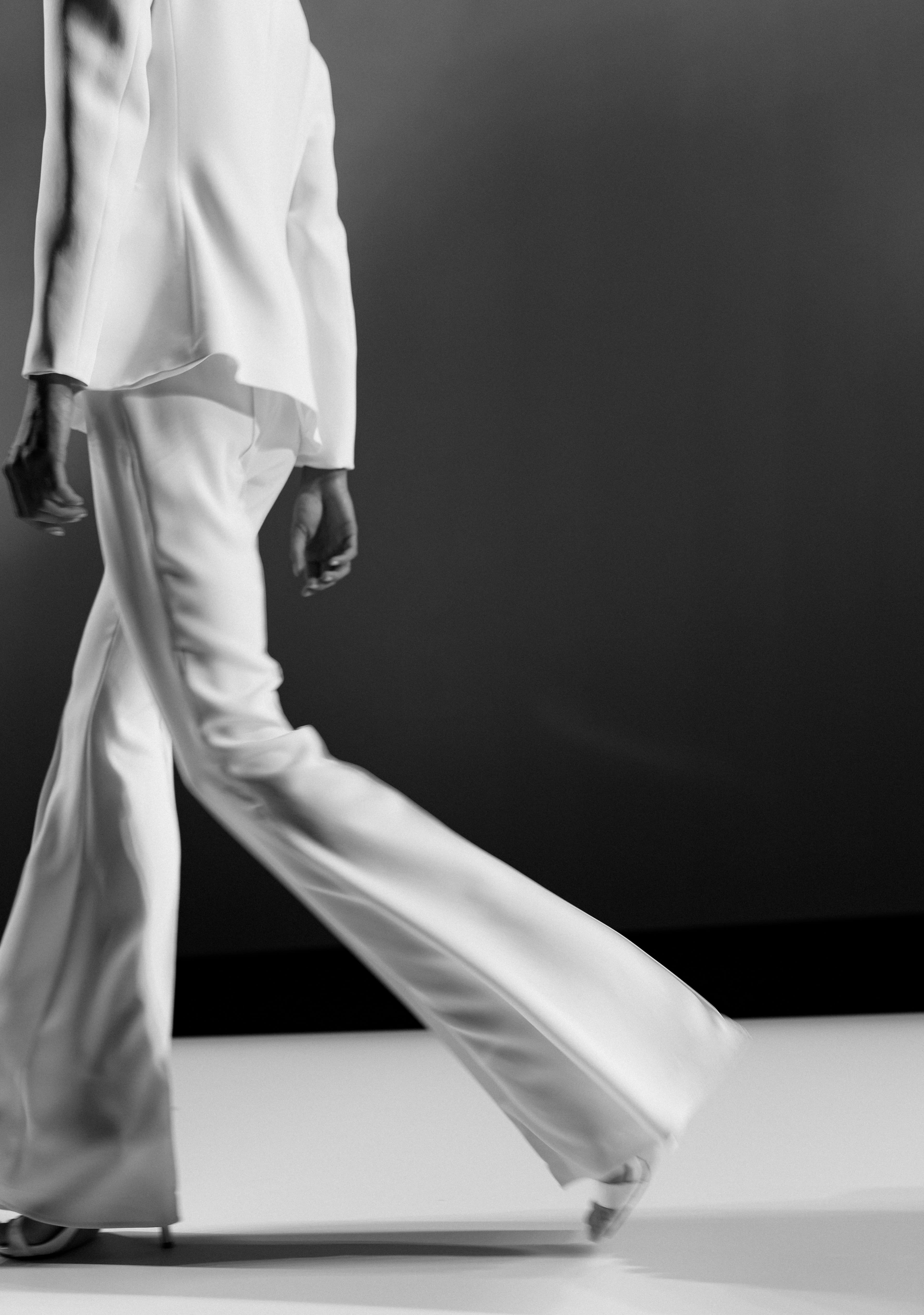 A person in a white suit strides forward with flowing wide leg trousers in monochrome.