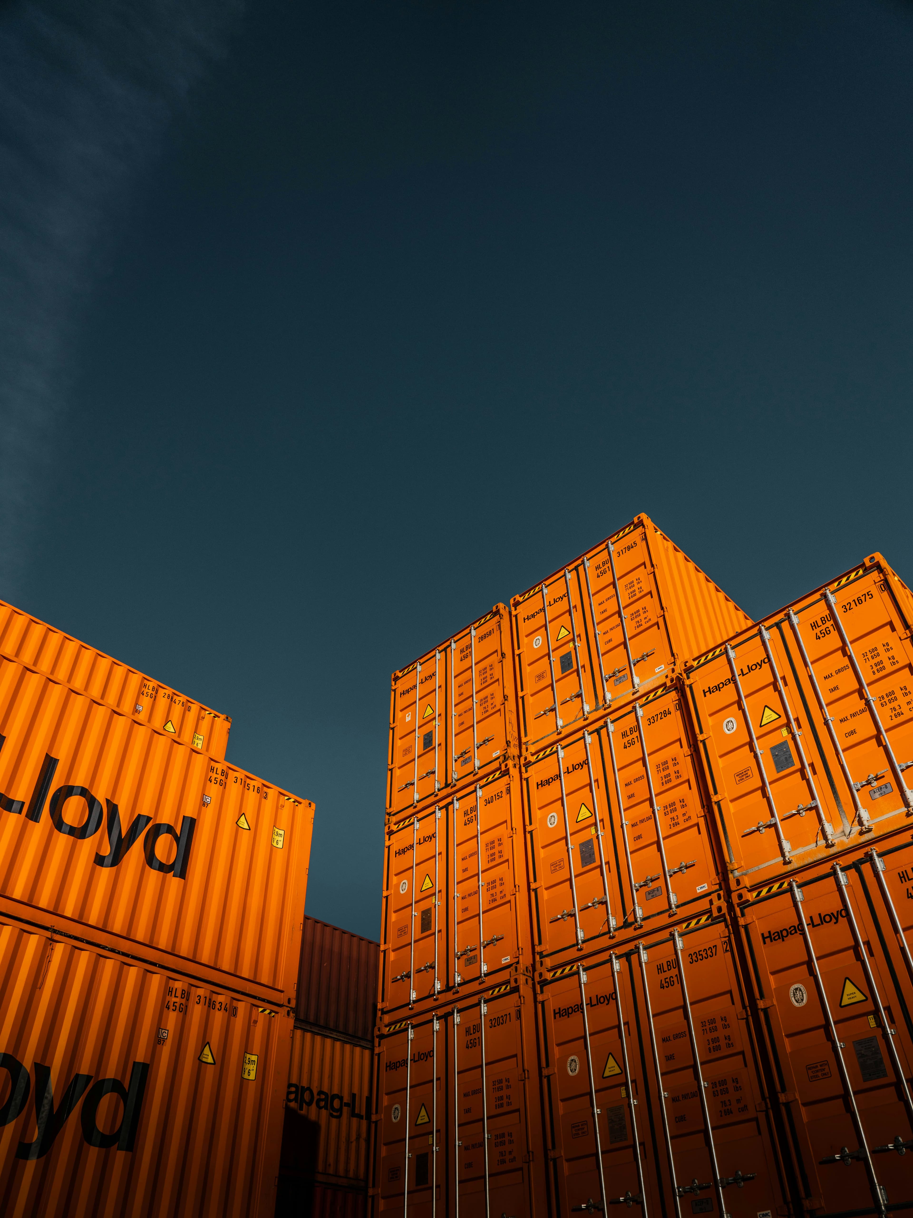 Stacks of bright orange Hapag-Lloyd shipping containers reaching toward a deep, dark blue sky.