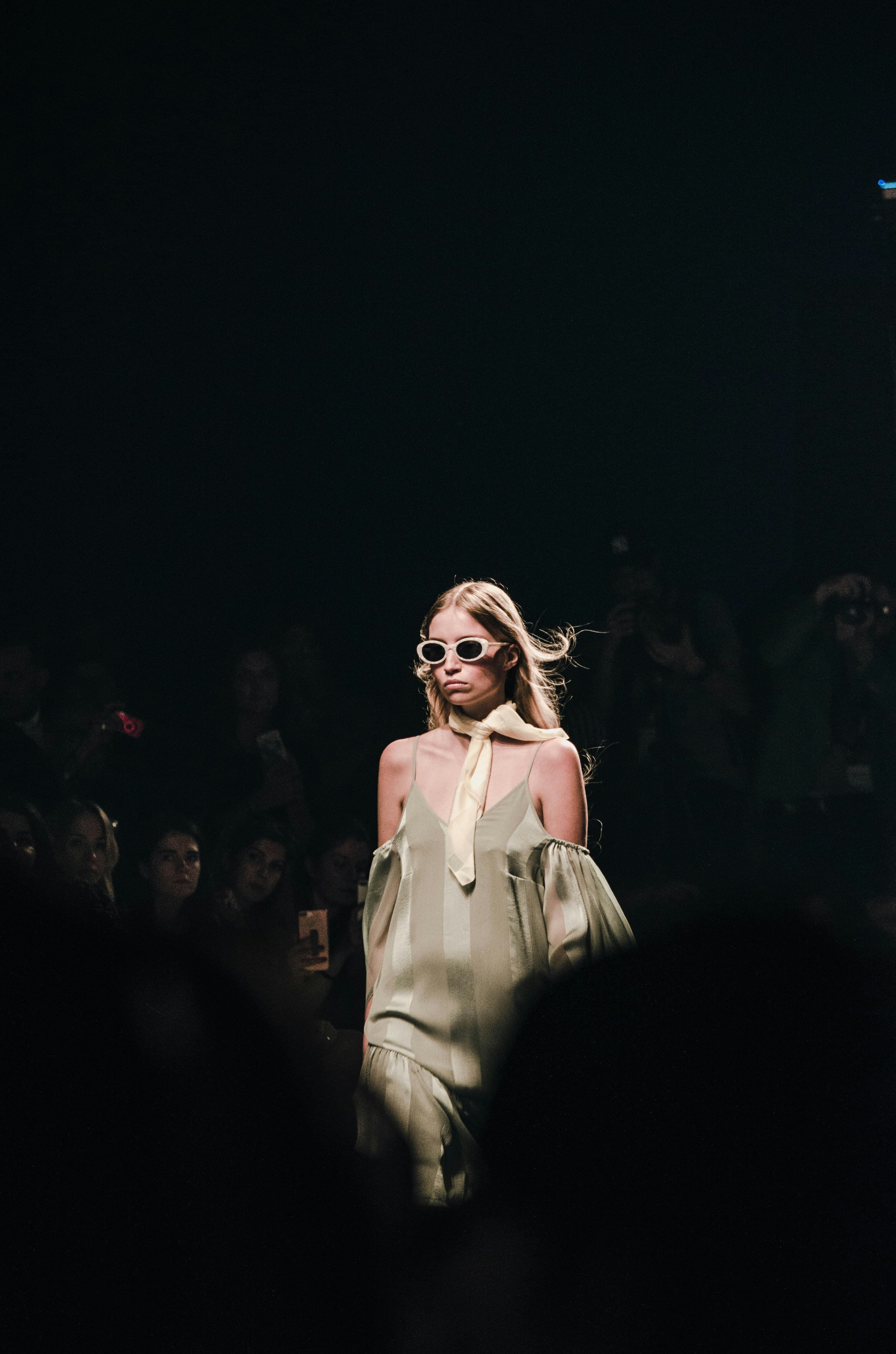 Blonde model in a green off-the-shoulder dress and white sunglasses walks a dark runway.
