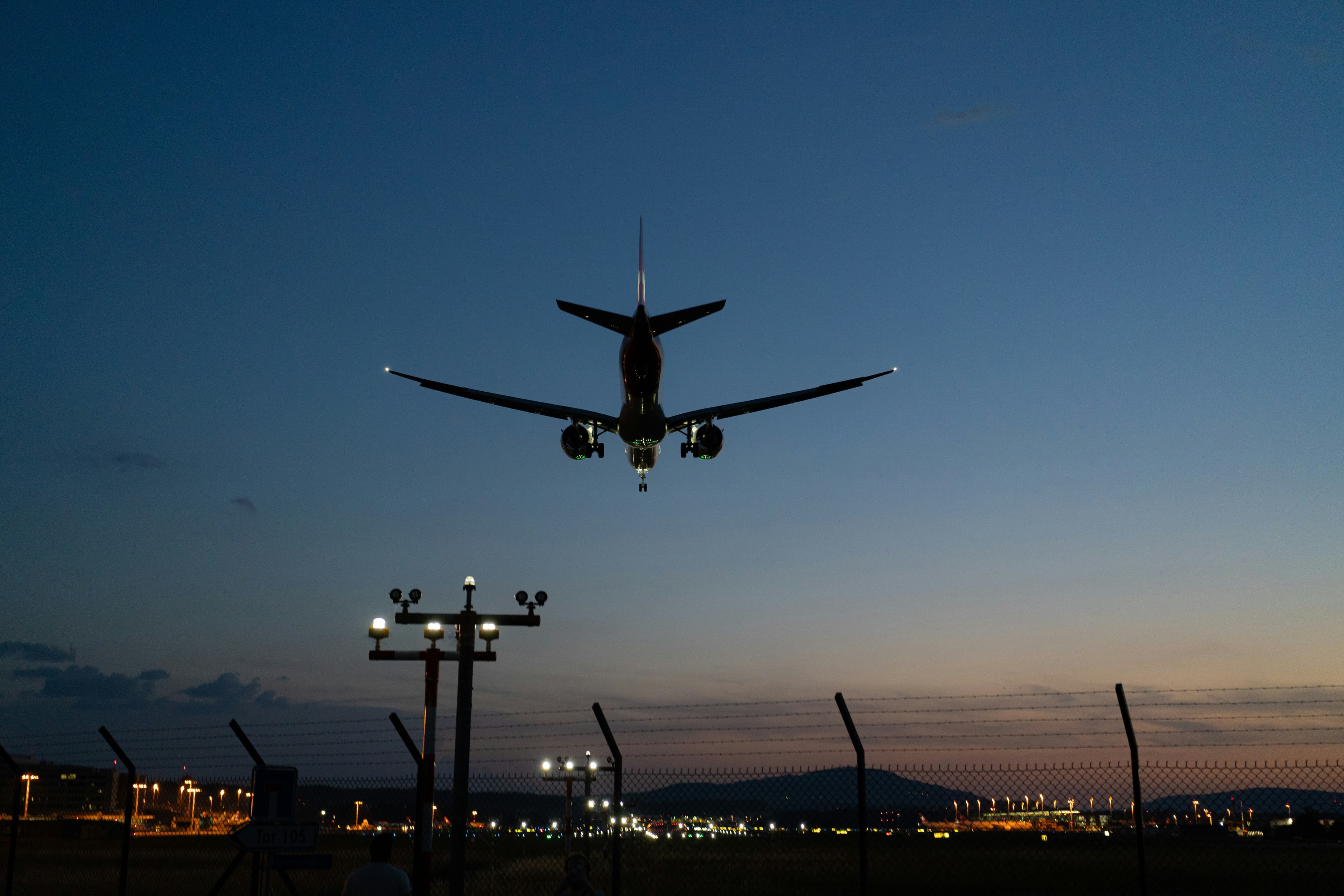 Silhouette of an airplane landing at dusk over an airport fence with runway lights.