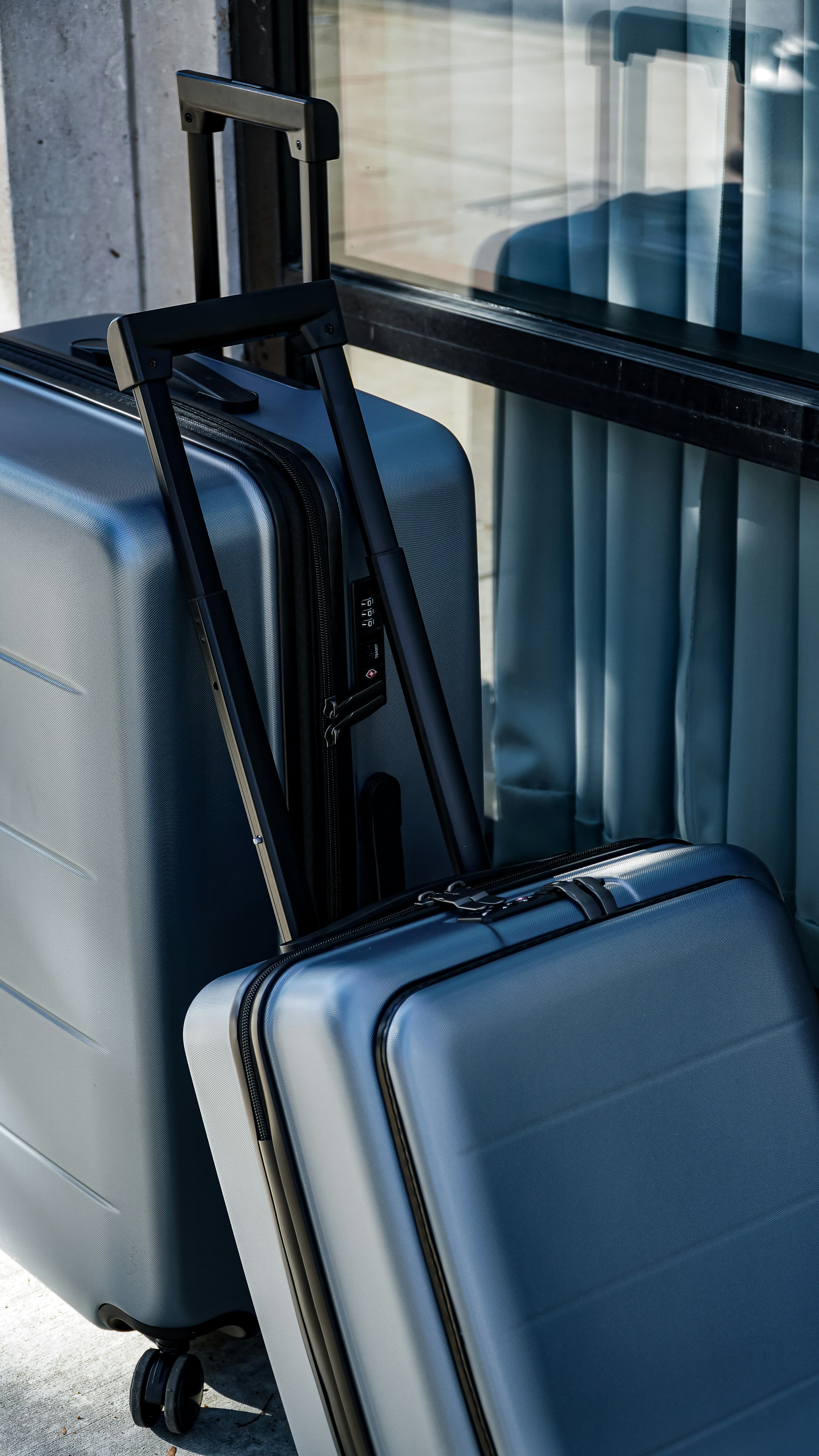 Two blue hardside suitcases with extended handles leaning against a glass window in bright sunlight.